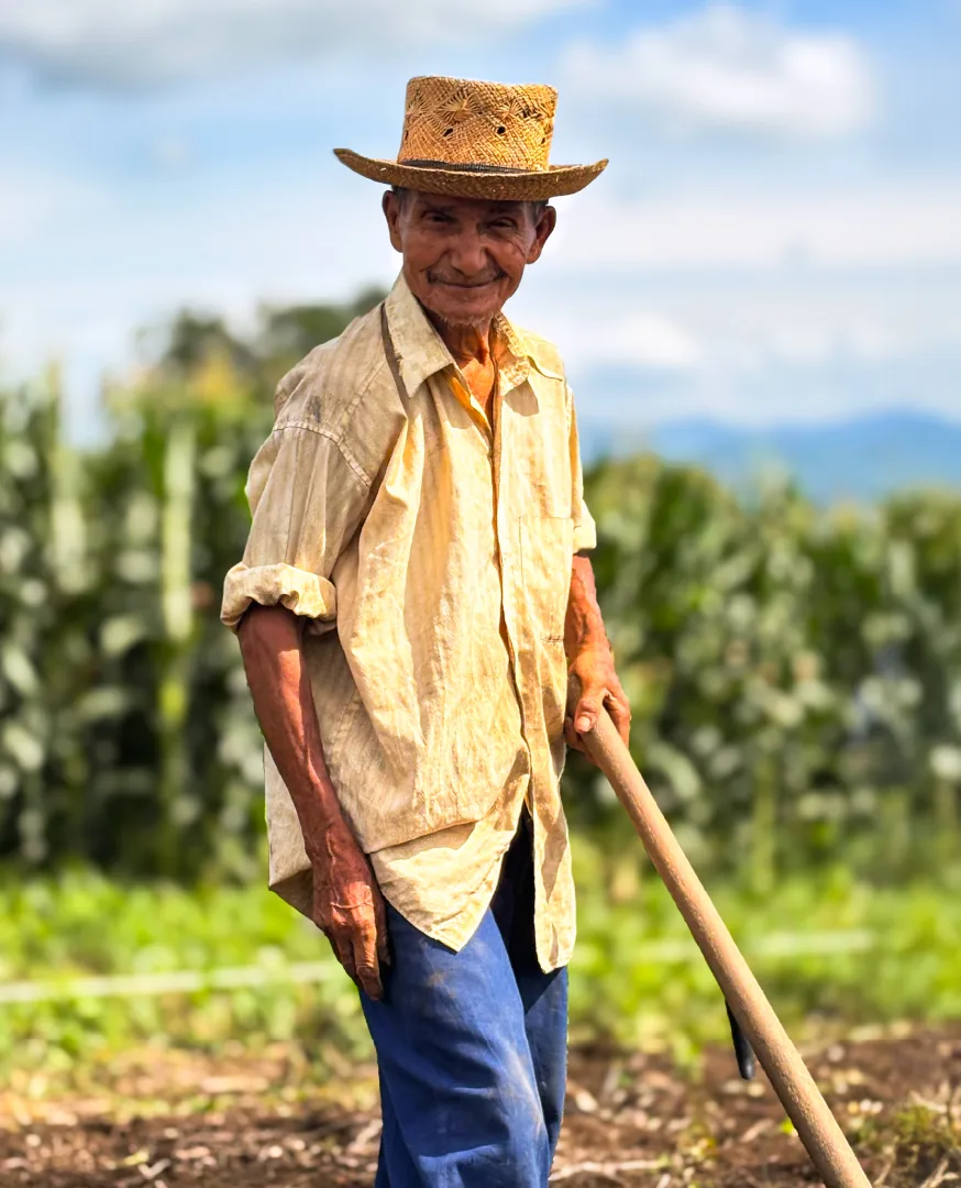 Pedro Ramírez, agricultor de sombrero de palma, sonríe apoyado en su herramienta en el campo